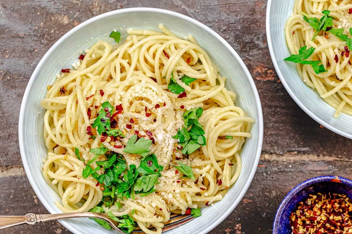 a close up of spaghetti aglio e olio topped with grated parmesan, red pepper flakes and parsley in a bowl with a fork next to another bowl of spaghetti aglio e olio and a small bowl of red pepper flakes.
