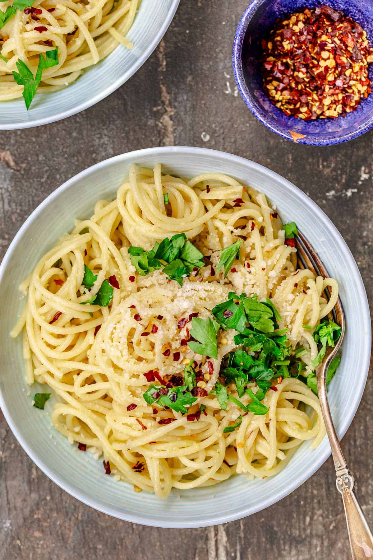 an overhead photo of spaghetti aglio e olio topped with grated parmesan, red pepper flakes and parsley in a bowl with a fork in front of another bowl of spaghetti aglio e olio and a small bowl of red pepper flakes.