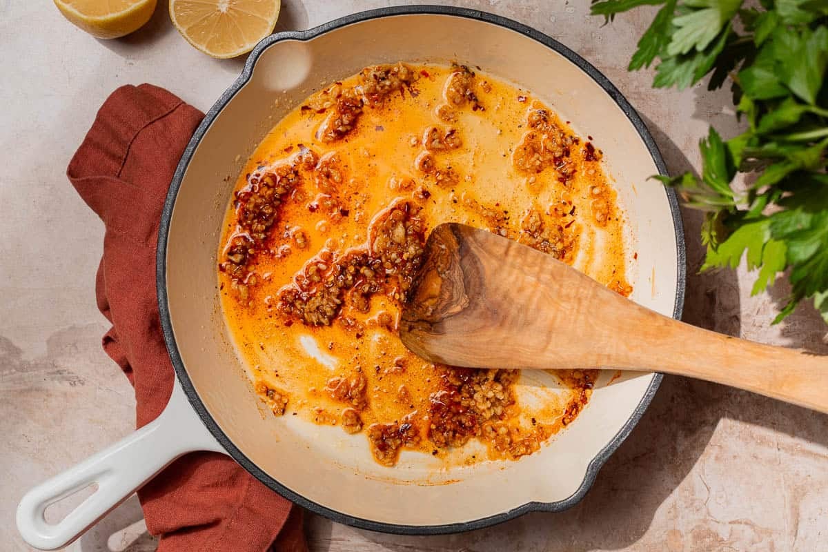 A white skillet with olive oil, red pepper flakes, Urfa pepper, cumin, paprika, garlic, salt and pepper being stirred with a wooden spoon.