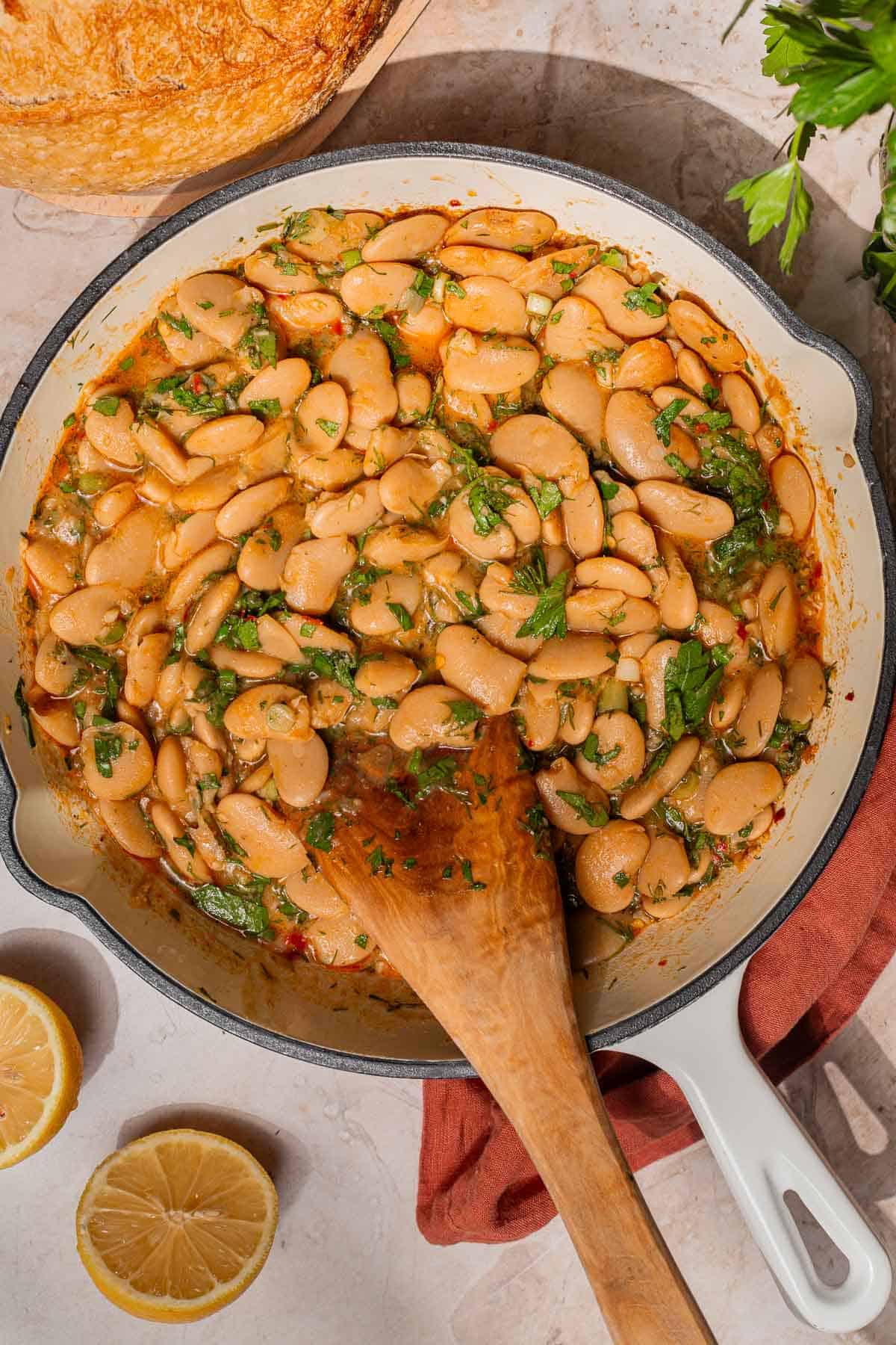 Butter beans in a white pan with a wooden spoon and bread and lemon wedges on the side.