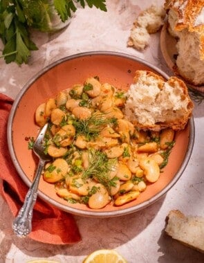 Serving of butter beans in a peach-colored bowl with a silver fork and a torn piece of crusty bread.