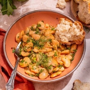 Serving of butter beans in a peach-colored bowl with a silver fork and a torn piece of crusty bread.