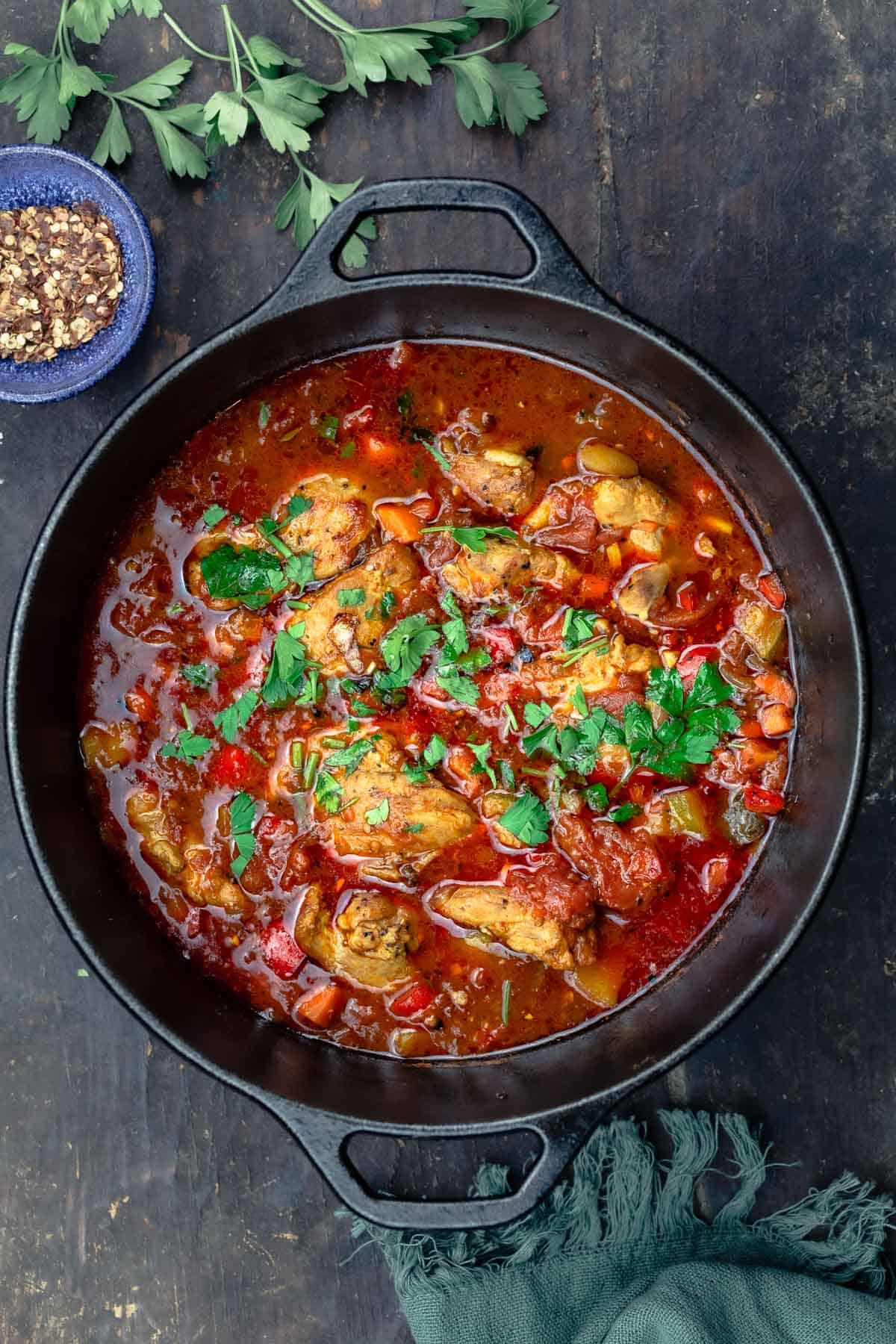 overhead photo of chicken stew garnished with parsley in a large pot next to extra parsley and a small bowl of red pepper flakes.