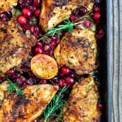a close up of baked cranberry chicken topped with fresh cranberries, sprigs of fresh rosemary and lemon slices in a cast iron pan.
