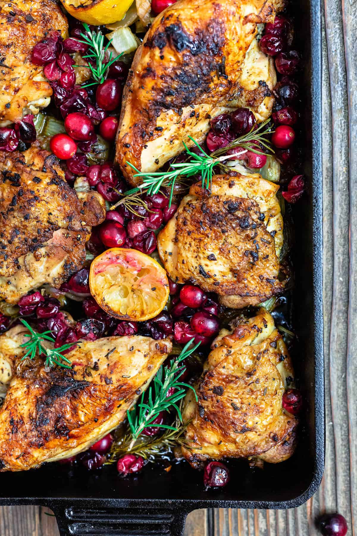 a close up of baked cranberry chicken topped with fresh cranberries, sprigs of fresh rosemary and lemon slices in a cast iron pan.