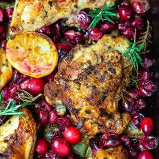 a close up of baked cranberry chicken topped with fresh cranberries, sprigs of fresh rosemary and lemon slices.