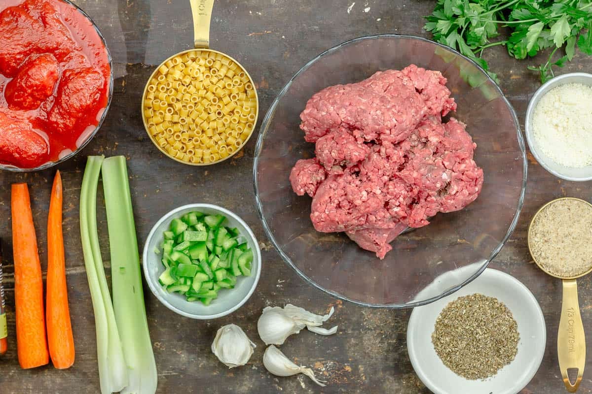 ingredients for meatball soup including ground beef, grated parmesan, bread crumbs, oregano, parsley, garlic, green pepper, canned whole tomatoes, carrots, celery, and ditalini pasta.