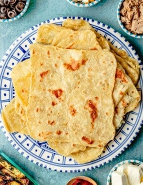 mlewi flatbreads stacked on a plate, surrounded by small bowls of olives, hard boiled eggs, tuna, cream cheese, harissa and a dish of homemade oven fries.