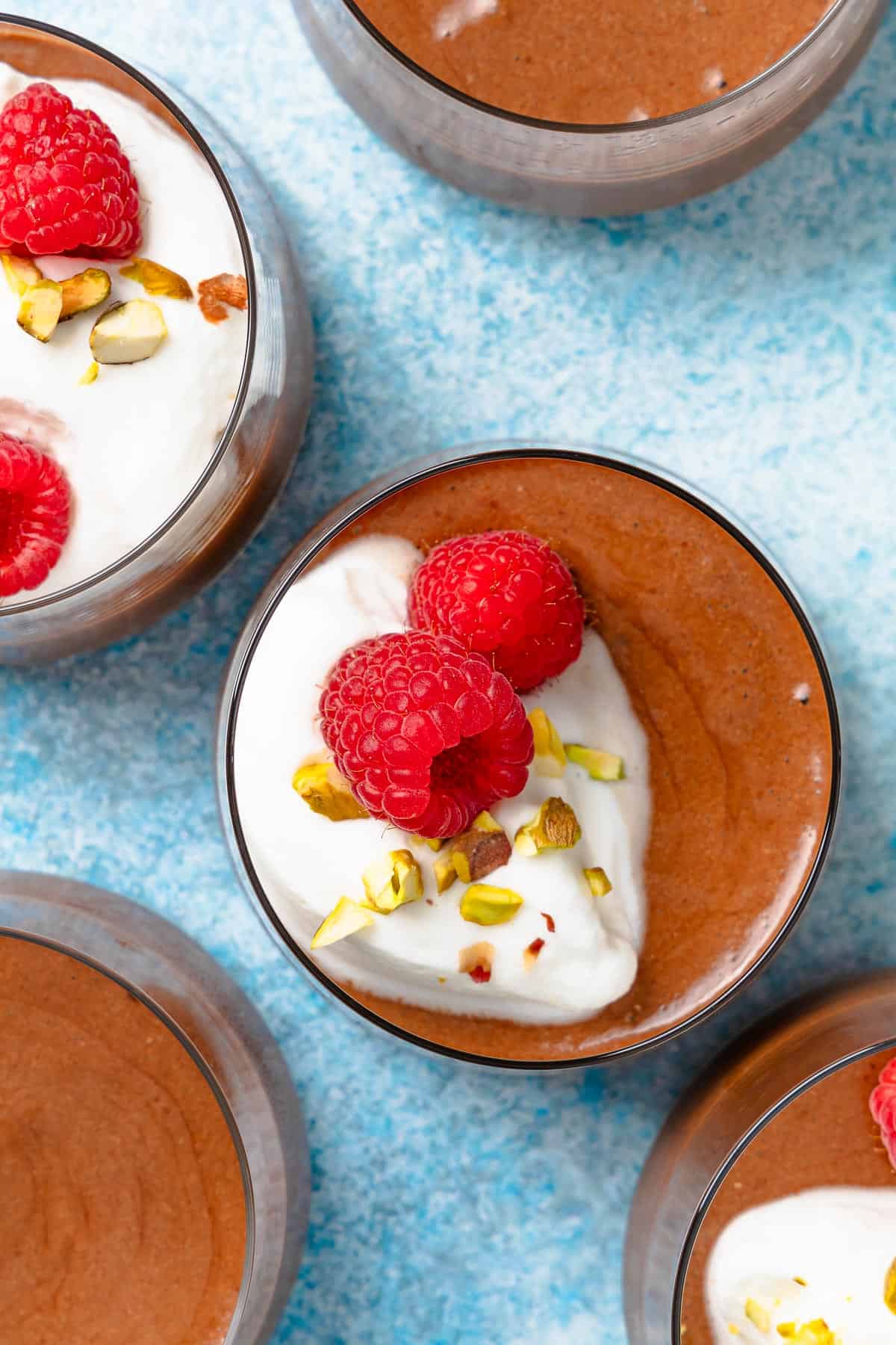 Overhead shot of chocolate mousse in glasses with whipped cream, crushed pistachios, and fresh raspberries on top.