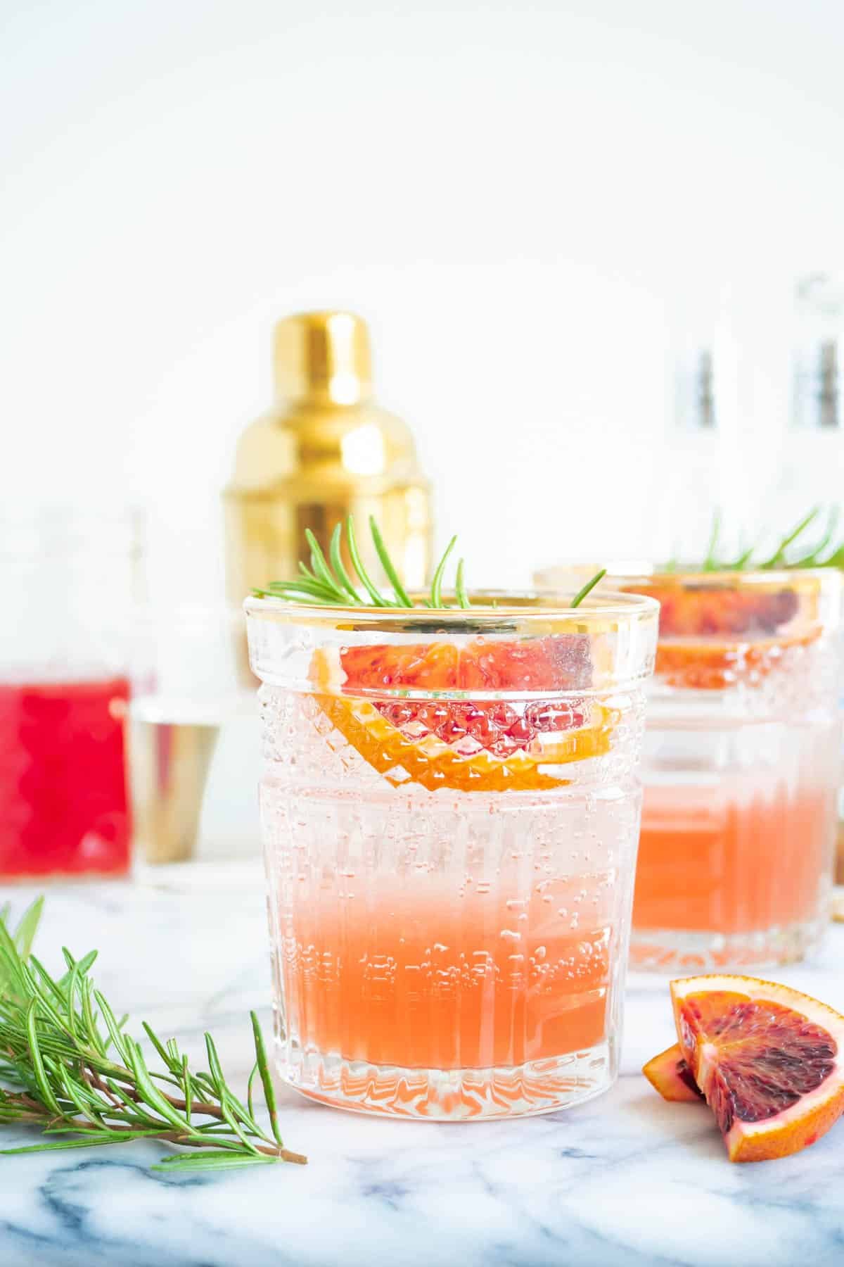 two blood orange rosemary cocktails garnished with sprigs of rosemary and wedges of blood orange next to another wedge of blood orange and a sprig of rosemary.