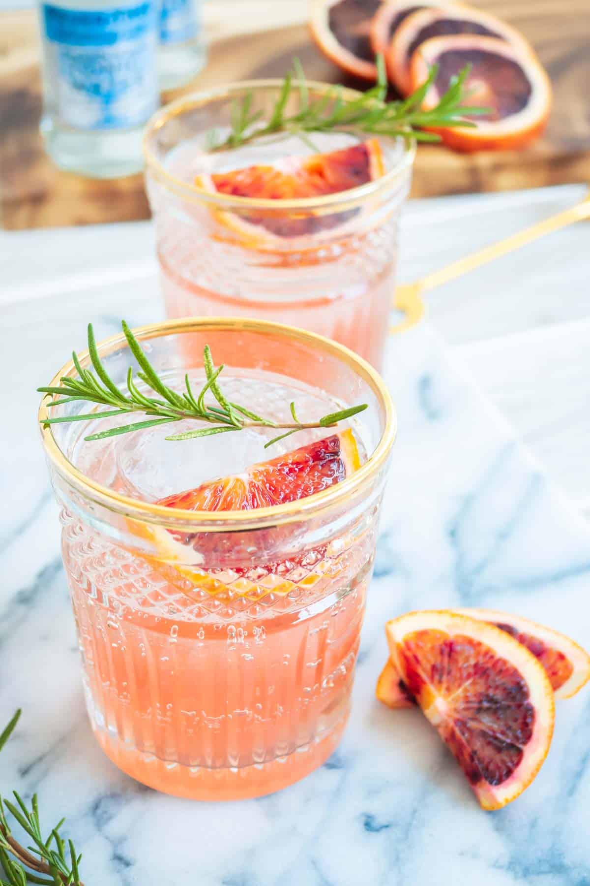 two blood orange rosemary cocktails garnished with sprigs of rosemary and wedges of blood orange next to more wedges of a blood orange.