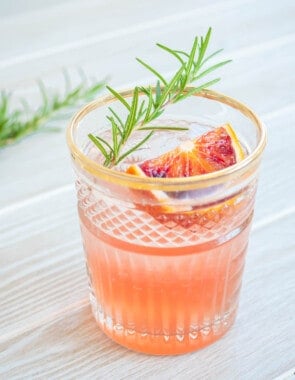 a blood orange rosemary cocktail garnished with a sprig of rosemary and a wedge of blood orange.