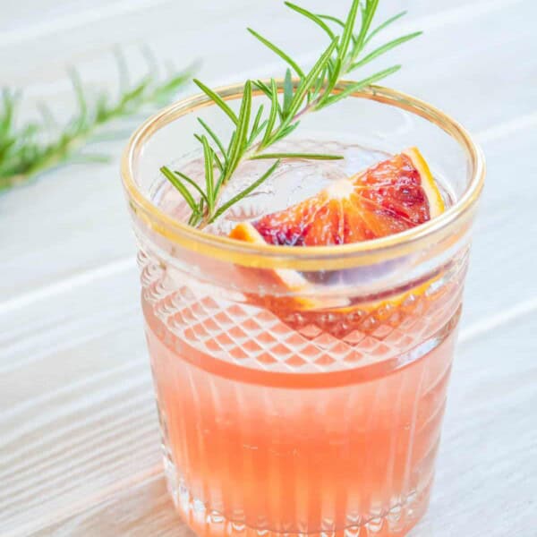 a blood orange rosemary cocktail garnished with a sprig of rosemary and a wedge of blood orange.