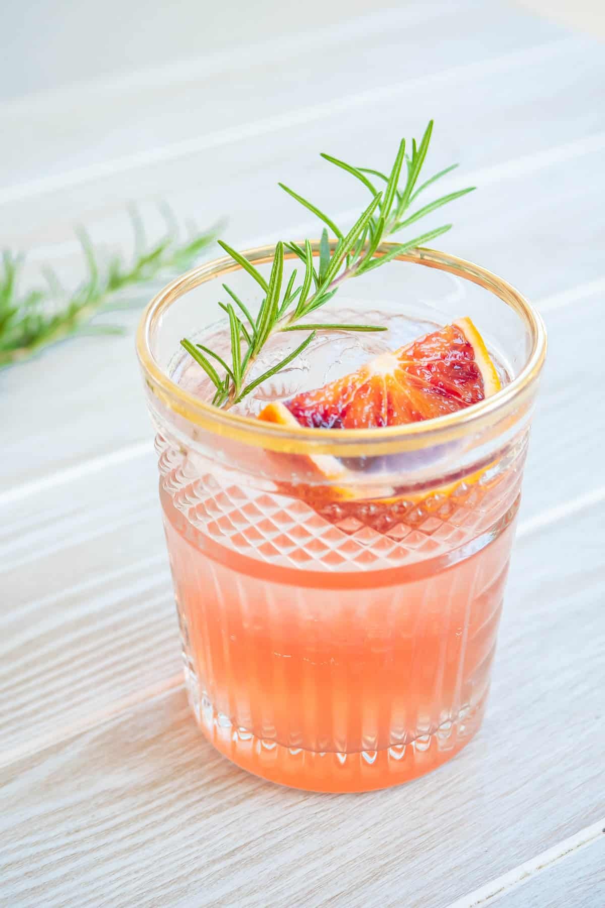 a blood orange rosemary cocktail garnished with a sprig of rosemary and a wedge of blood orange.