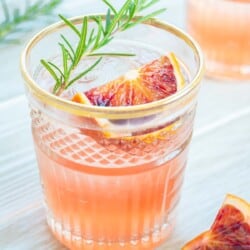 a blood orange rosemary cocktail garnished with a sprig of rosemary and a wedge of blood orange next to another wedge of blood orange.