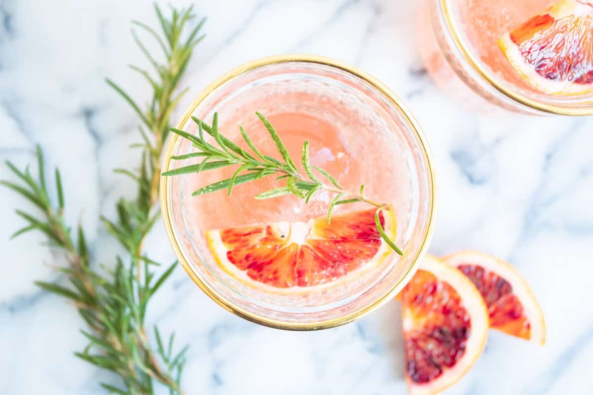 an overhead photo of a blood orange rosemary cocktail garnished with a sprig of rosemary and a wedge of blood orange next to another wedge of blood orange and more sprigs of rosemary.