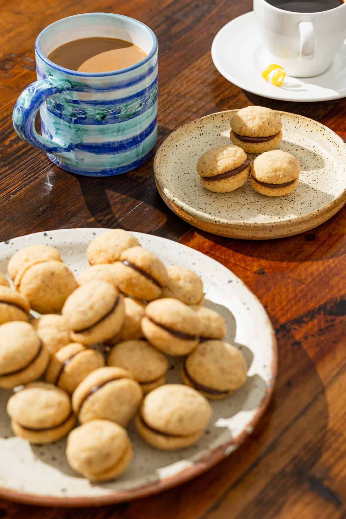 a serving plate with baci di dama hazelnut cookies next to a plate with three cookies and two cups of coffee.