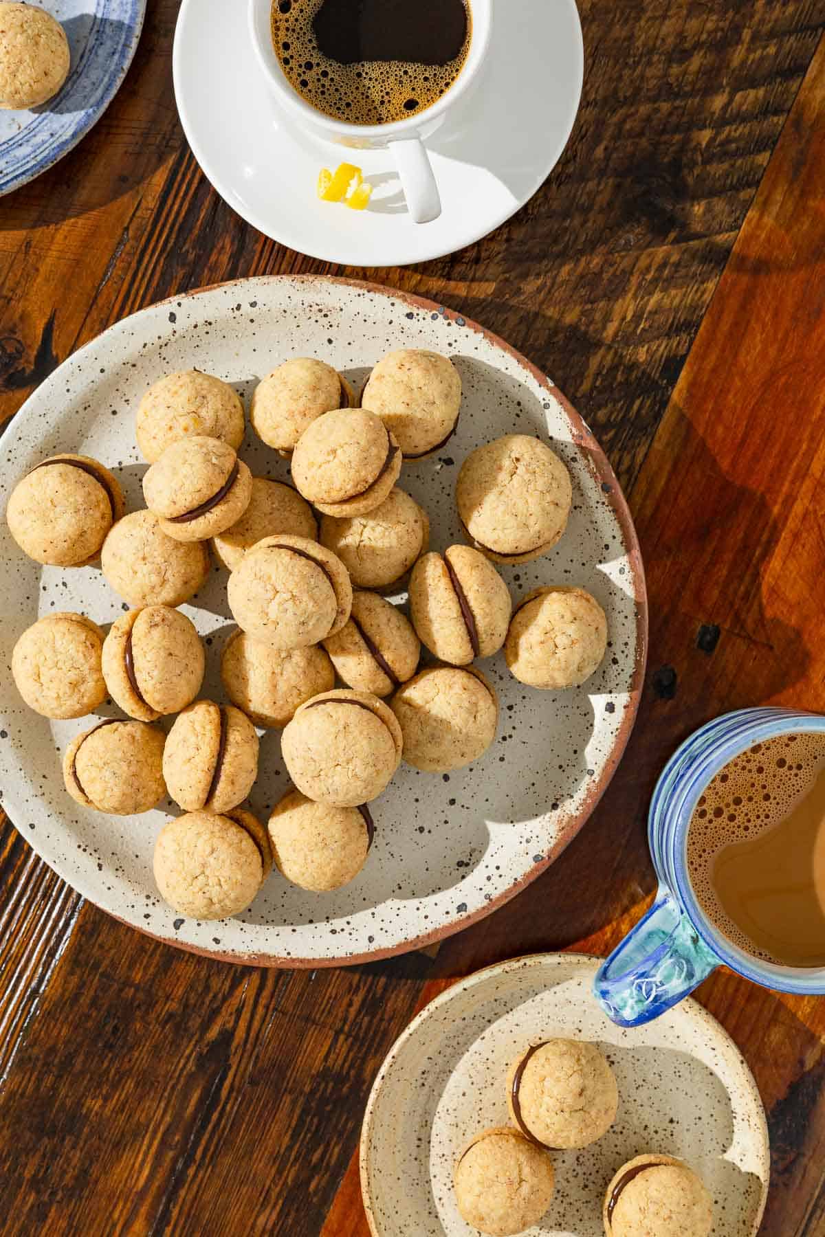 an overhead photo of a serving plate with baci di dama hazelnut cookies next to a plate with three cookies and two cups of coffee.