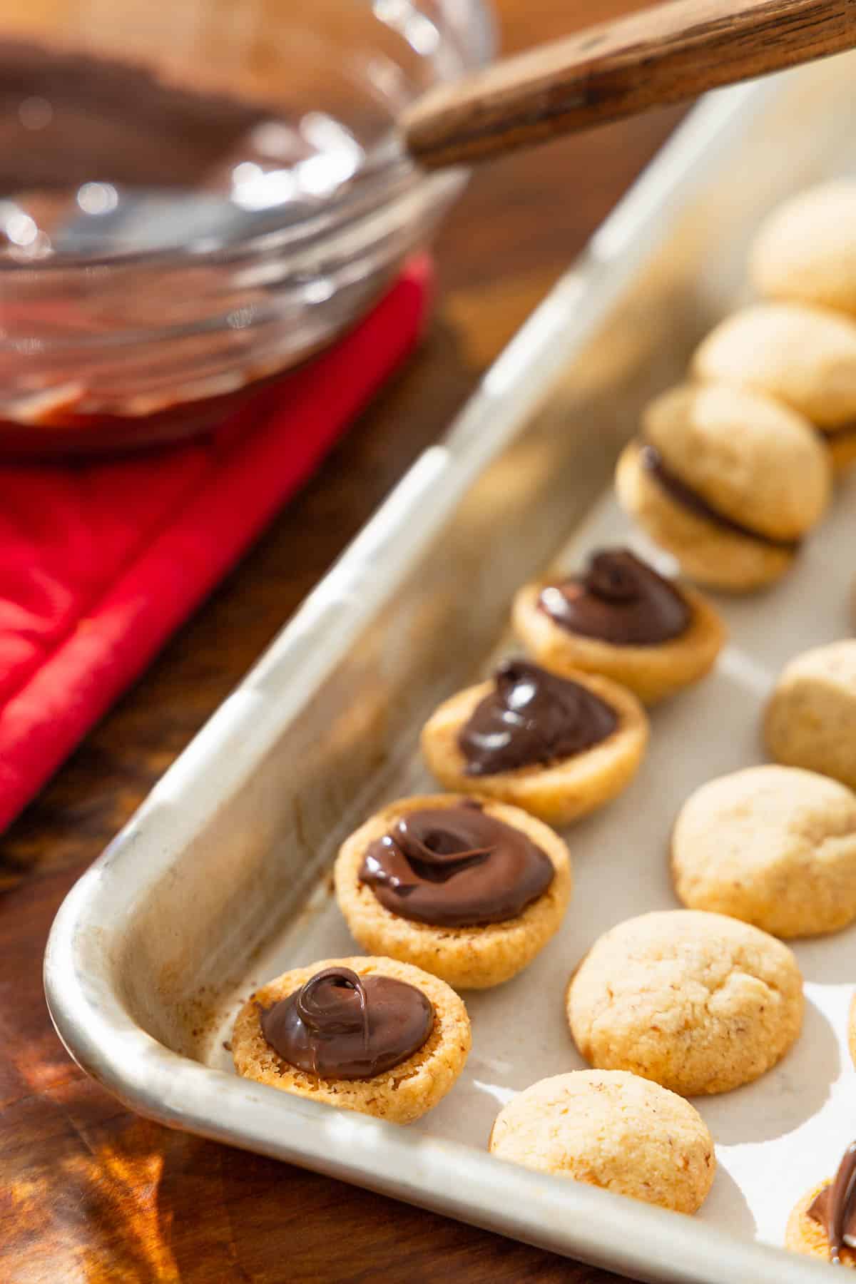 baci di dama hazelnut cookies on a baking sheet being filled with melted chocolate to make them sandwich cookies.