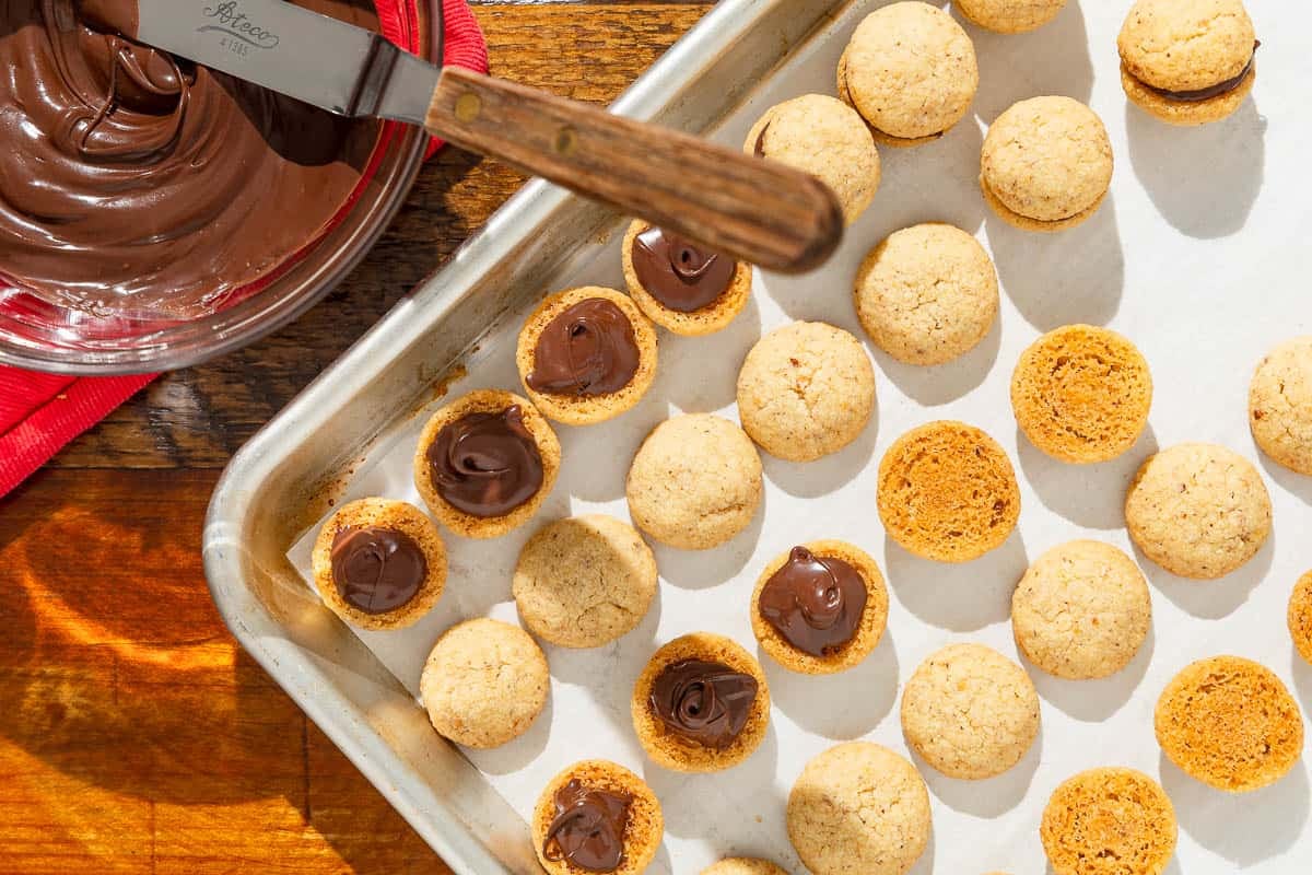baci di dama hazelnut cookies on a baking sheet being filled with melted chocolate to make them sandwich cookies.