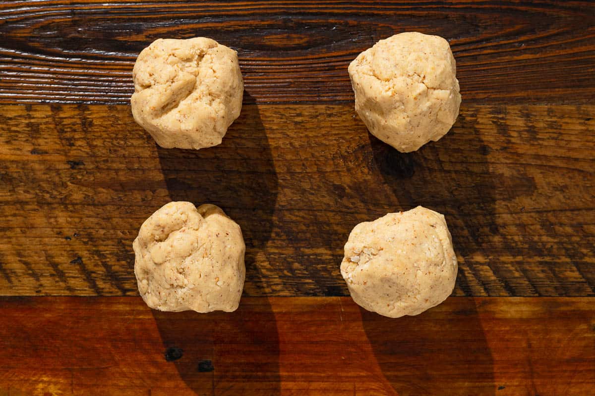 four equal portions of baci di dama hazelnut cookie dough rolled into balls on a table.