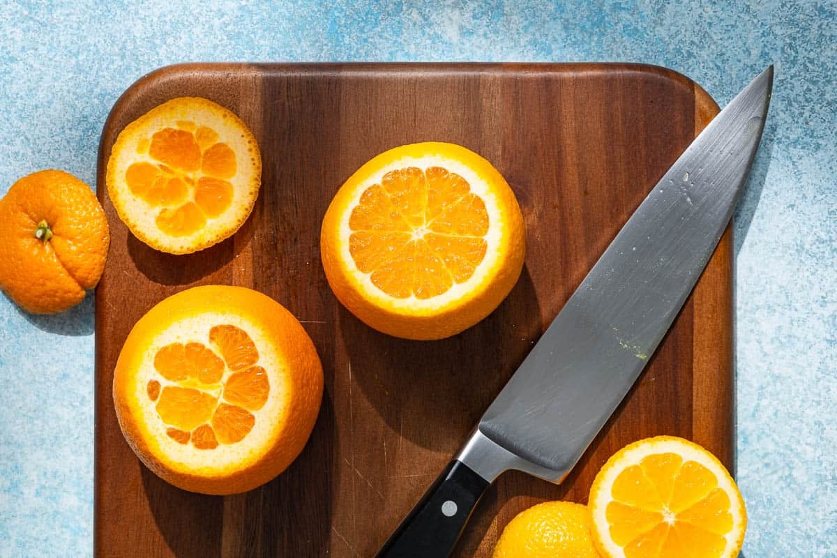 two oranges with their tops and bottoms cut off with a knife on a wooden cutting board.