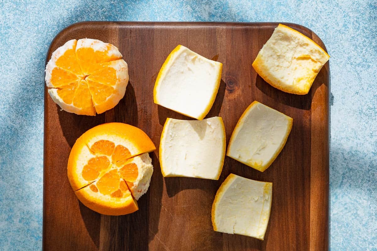 two oranges on a cutting board, one with its peel totally cut off and one with only one piece of peel cut off, next to 5 pieces of orange peels.