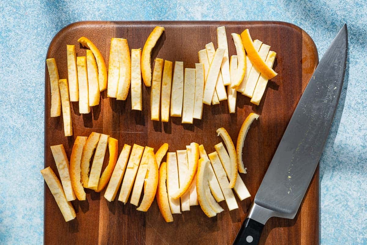 an overhead photo of orange peels just after being sliced into quarter inch wide pieces with a knife on a cutting board.