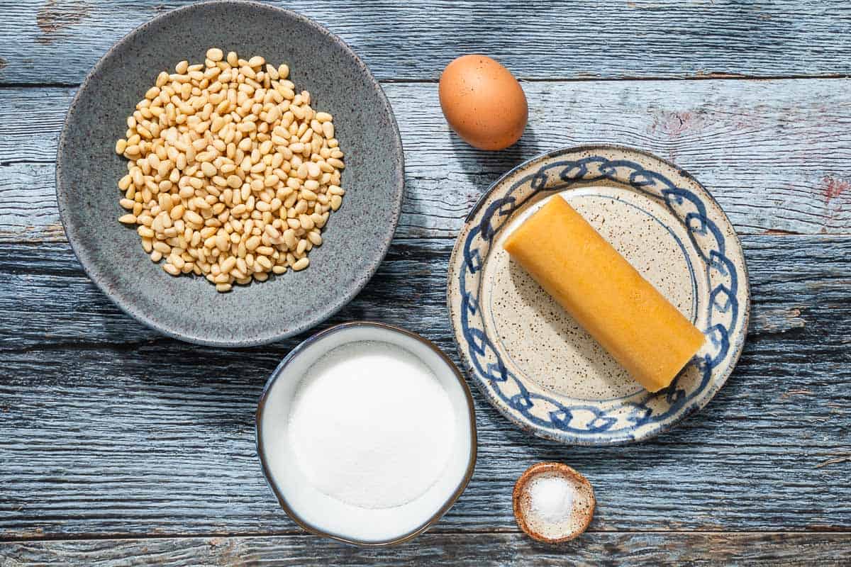 ingredients for pignoli cookies including almond paste, sugar, egg, kosher salt and pine nuts.