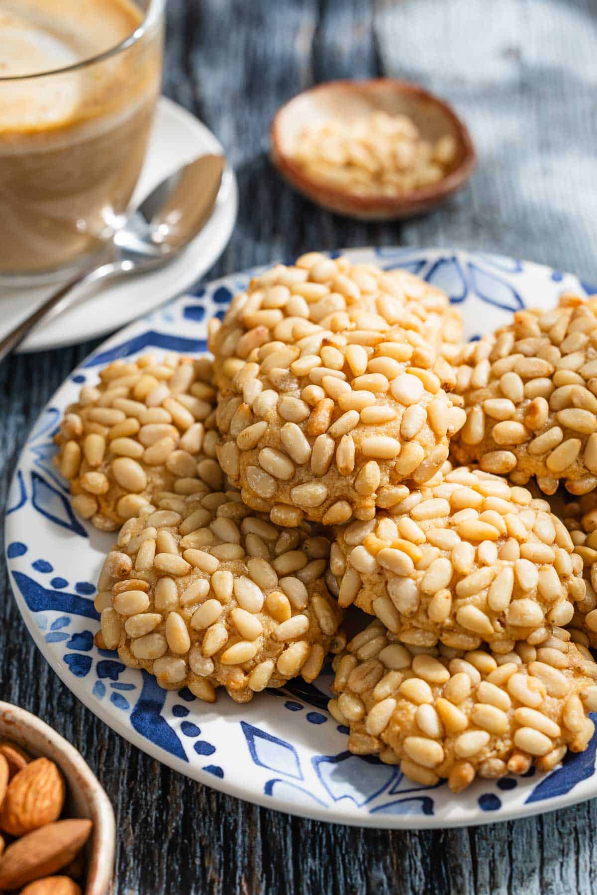 several pignoli cookies stacked on a serving plate next to a small bowl of pine nuts, a cup of coffee with a spoon, and a bowl of almonds.