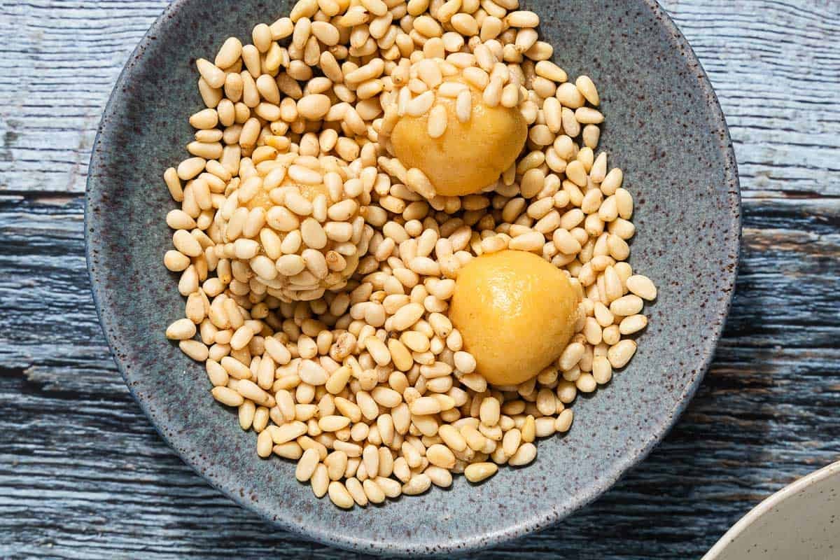 small pignoli cookie dough balls being rolled in pine nuts in a bowl.