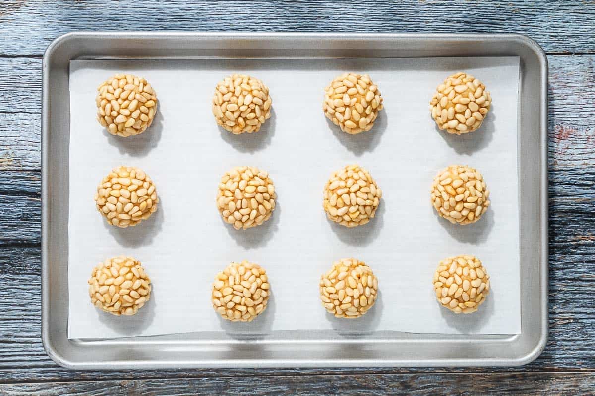 an overhead photo of 12 unbaked pignoli cookies spread out on a parchment lined baking sheet.