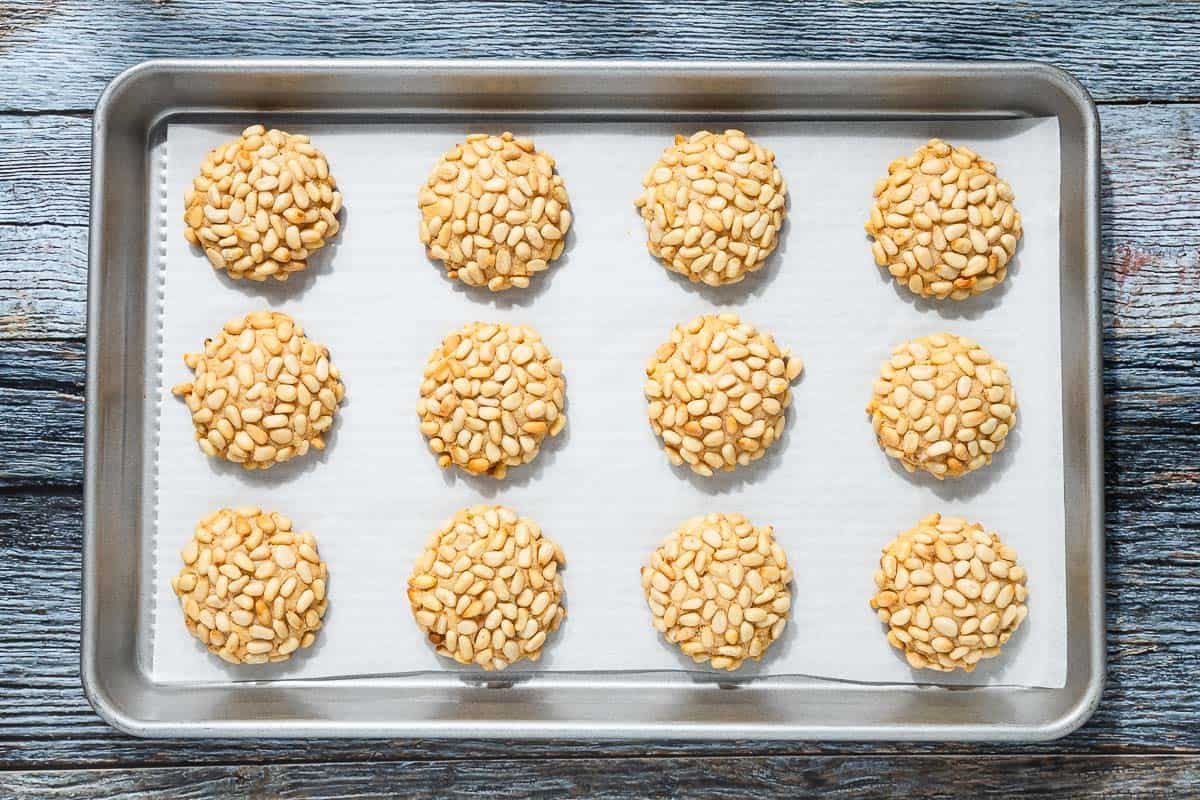 an overhead photo of 12 baked pignoli cookies spread out on a parchment lined baking sheet.