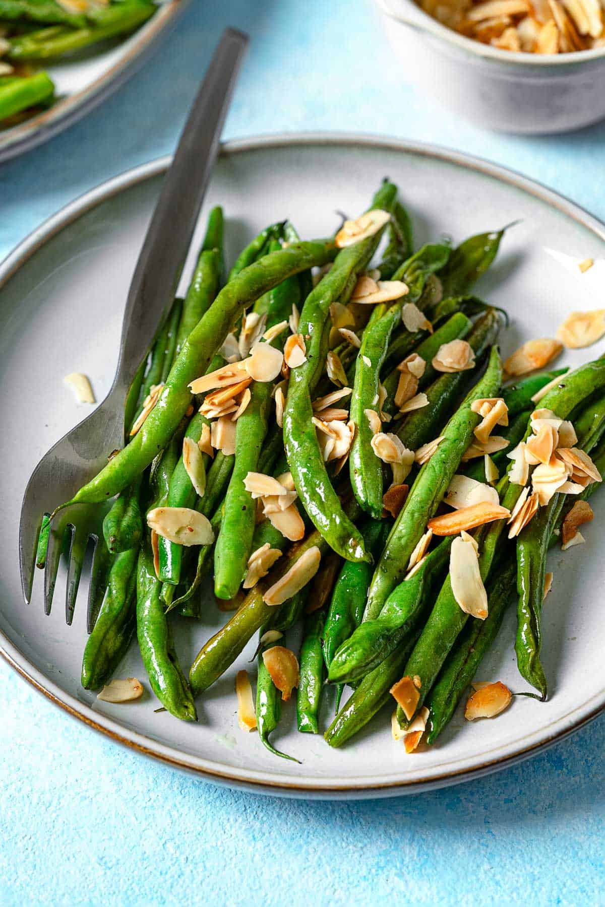 a close up of a serving of roasted green beans garnished with sliced almonds on a plate with a fork.