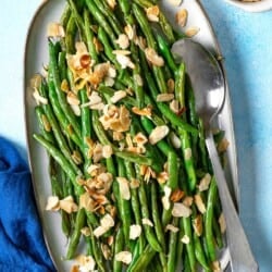 an overhead photo of a serving platter of roasted green beans garnished with sliced almonds with a serving spoon next to a bowl of sliced almonds.