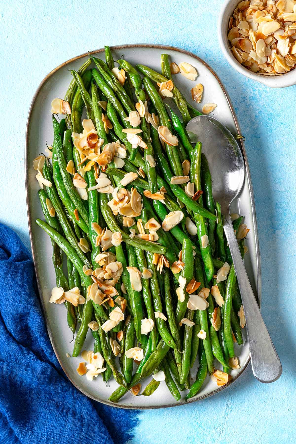 an overhead photo of a serving platter of roasted green beans garnished with sliced almonds with a serving spoon next to a bowl of sliced almonds.