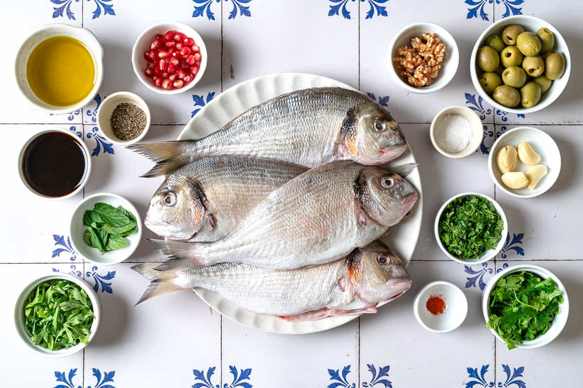 ingredients for persian baked fish including whole sea bream fish, saffron, salt, pepper, walnuts, garlic cloves, cilantro, parsley, mint, green olives, pomegranate molasses, olive oil and pomegranate seeds.