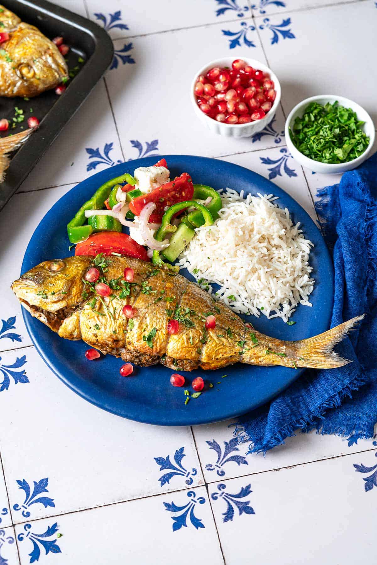 A serving of persian baked fish on a plate with rice and a salad next to small bowls of chopped parsley and pomegranate seeds and the baking sheet with the rest of the fish.