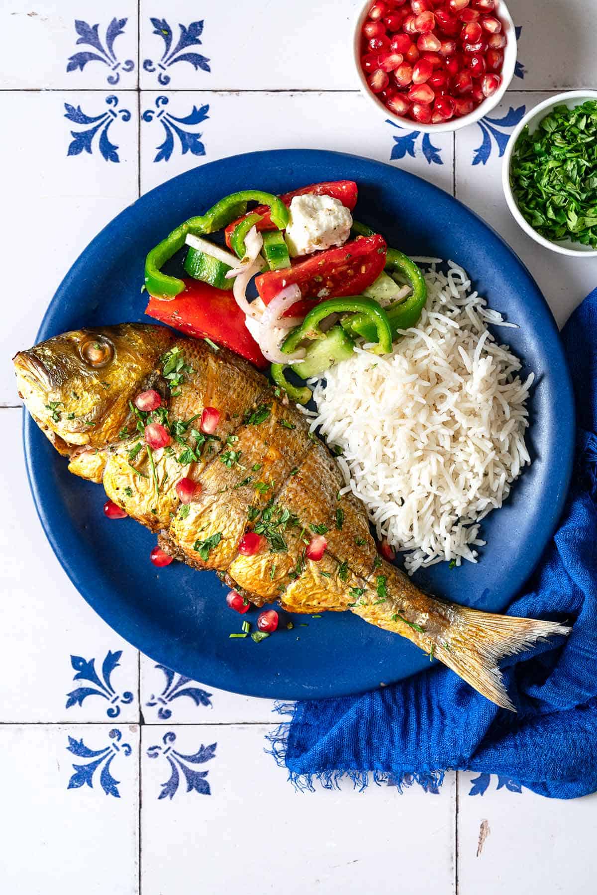 A serving of persian baked fish on a plate with rice and a salad next to small bowls of pomegranate seeds and chopped parsley.