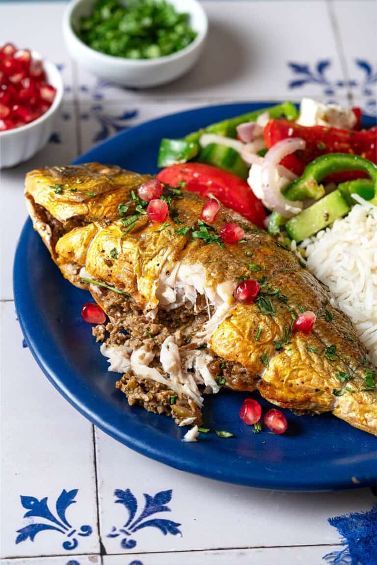 A serving of persian baked fish on a plate with rice and a salad. A part of the fish has been cut open showing the stuffing inside.