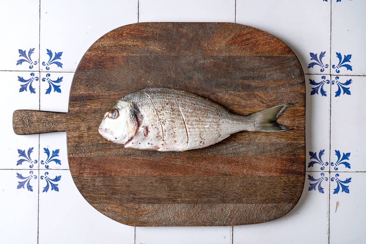 1 whole uncooked sea bream with slits cut into it laying on a cutting board.