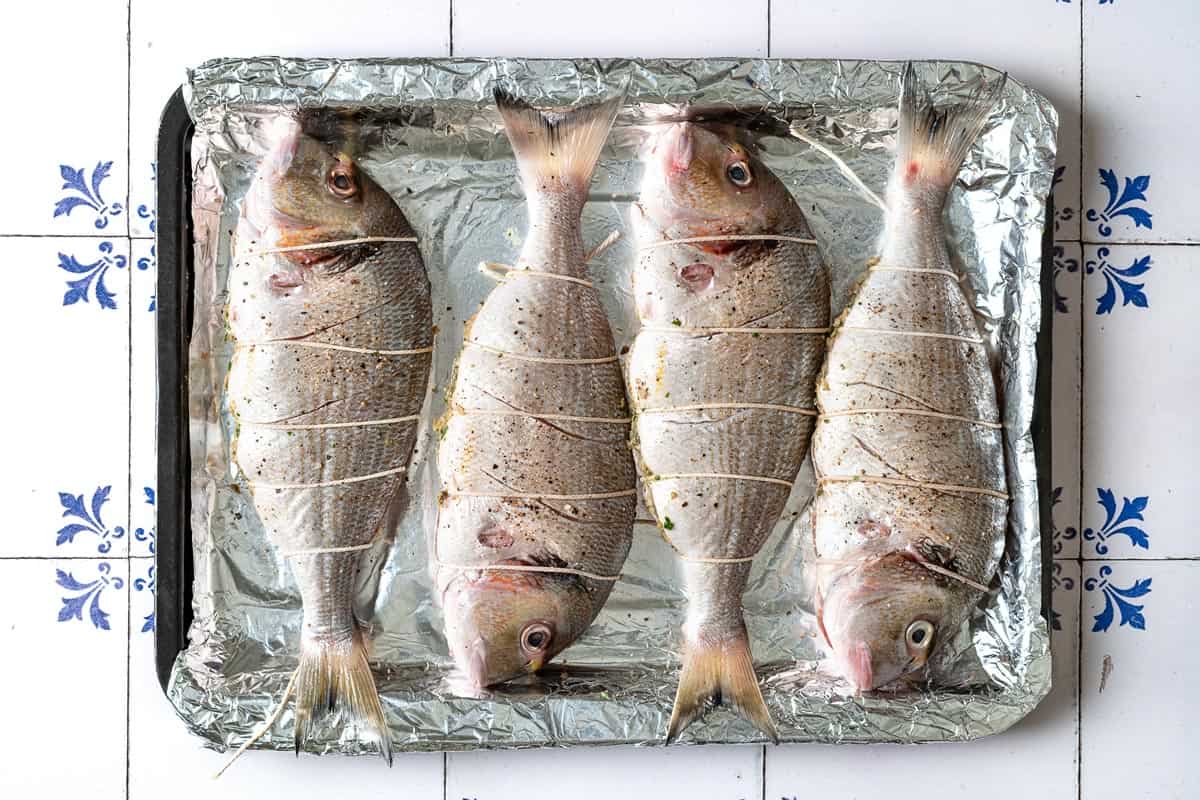 4 whole sea breams, stuffed and tied closed laying side by side on a foil lined baking sheet.