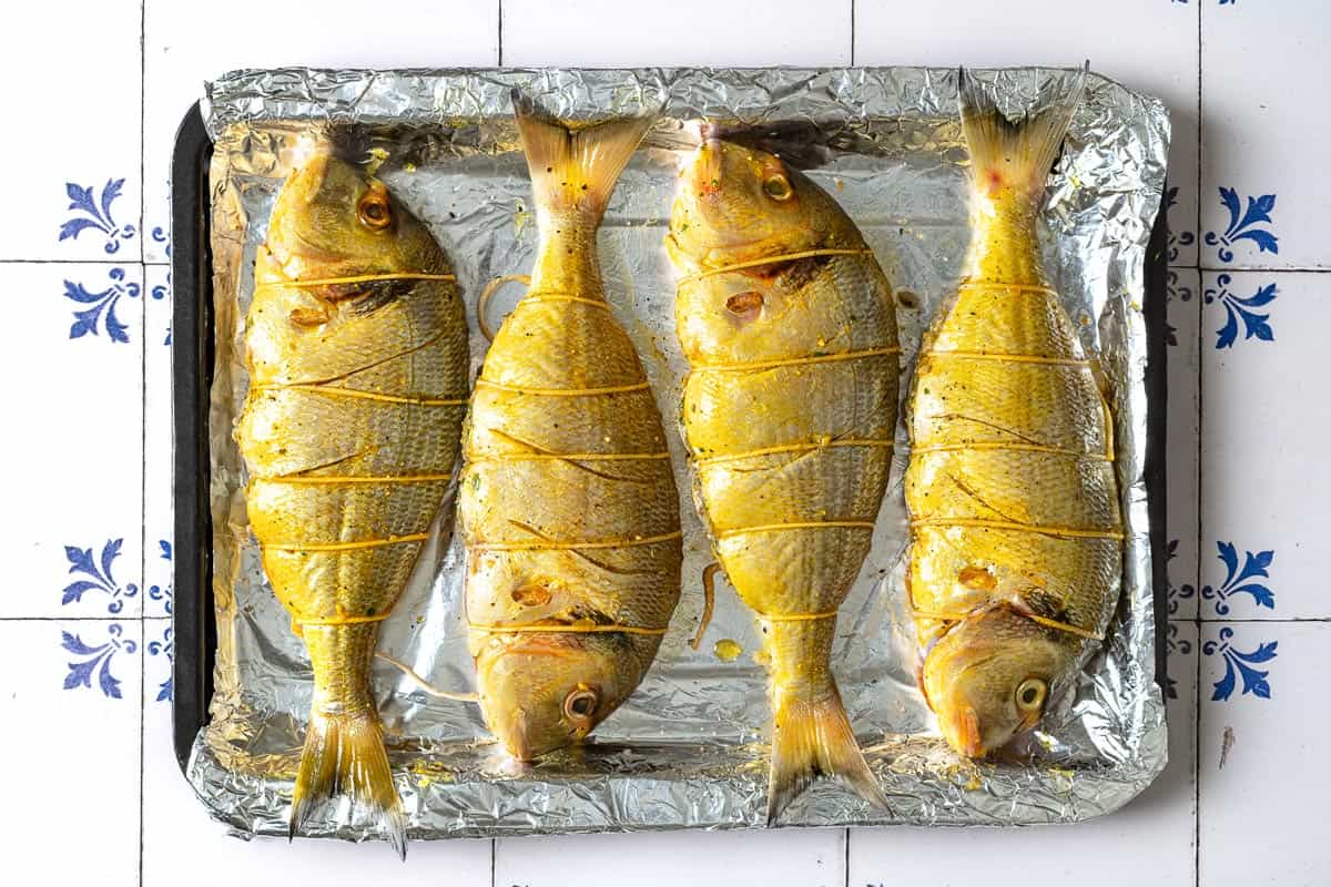 4 whole uncooked, stuffed sea breams covered in a saffron glaze, tied closed laying side by side on a foil lined baking sheet.