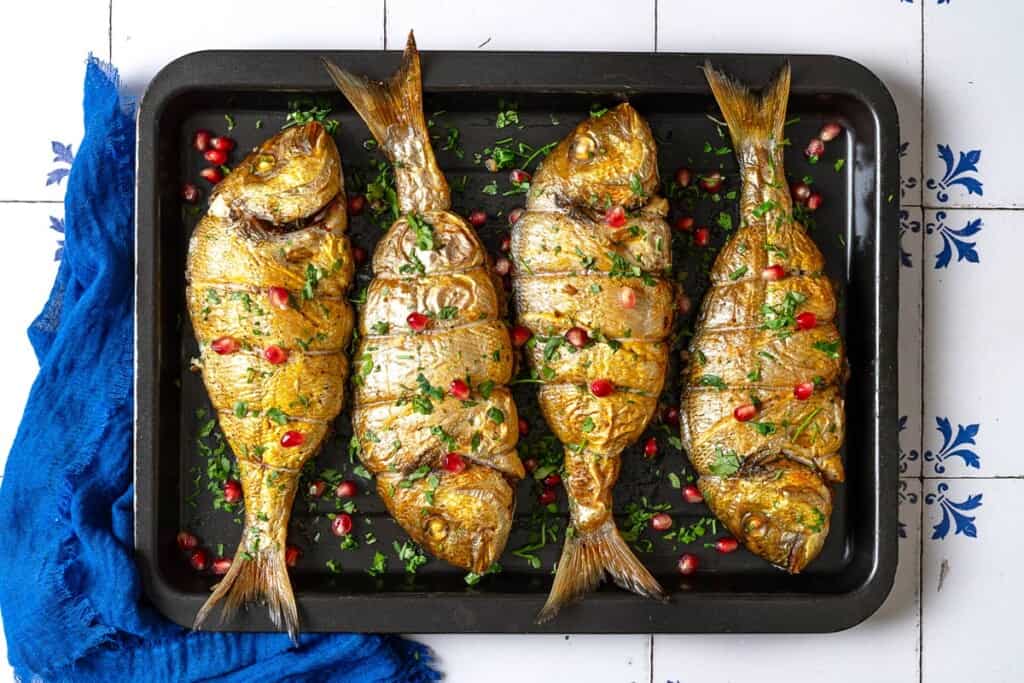 an overhead photo of 4 cooked persian baked fishes garnished with pomegranate seeds and parsley on a sheet pan.