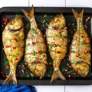 an overhead photo of 4 cooked persian baked fishes garnished with pomegranate seeds and parsley on a sheet pan.