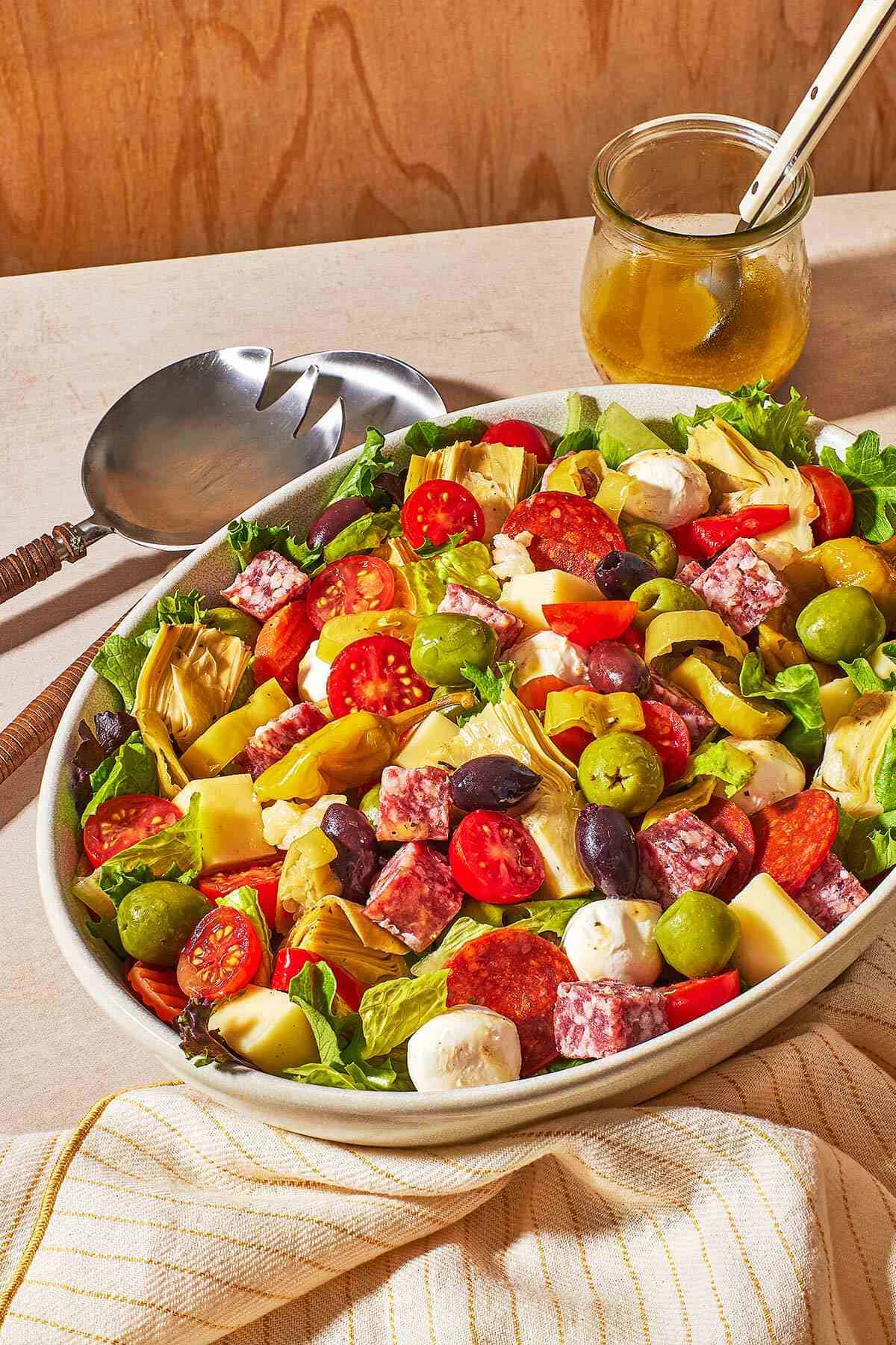 antipasto salad in a large serving bowl next to two serving utensils, dijon vinaigrette in a jar with a spoon, and a linen napkin.