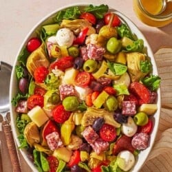 an overhead photo of antipasto salad in a serving bowl next to a jar of dijon dressing with a spoon, serving utensils and a linen napkin.