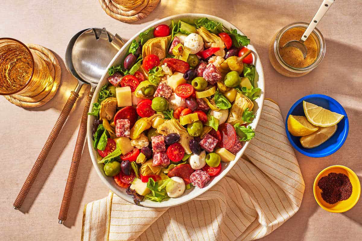 antipasto salad in a large serving serving bowl surrounded by serving utensils, a glass of water, lemon wedges, sumac and a jar of dijon vinaigrette with a spoon.
