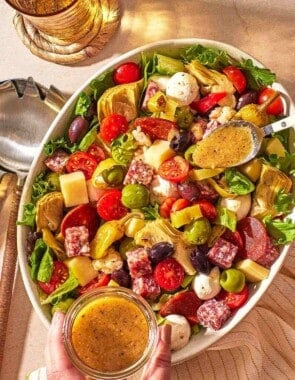 an overhead photo of dijon vinaigrette being spooned from a jar onto the antipasto salad, next to a glass of water and serving utensils.