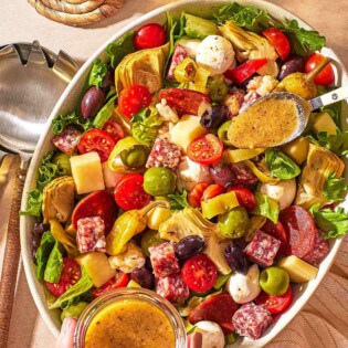 an overhead photo of dijon vinaigrette being spooned from a jar onto the antipasto salad, next to a glass of water and serving utensils.